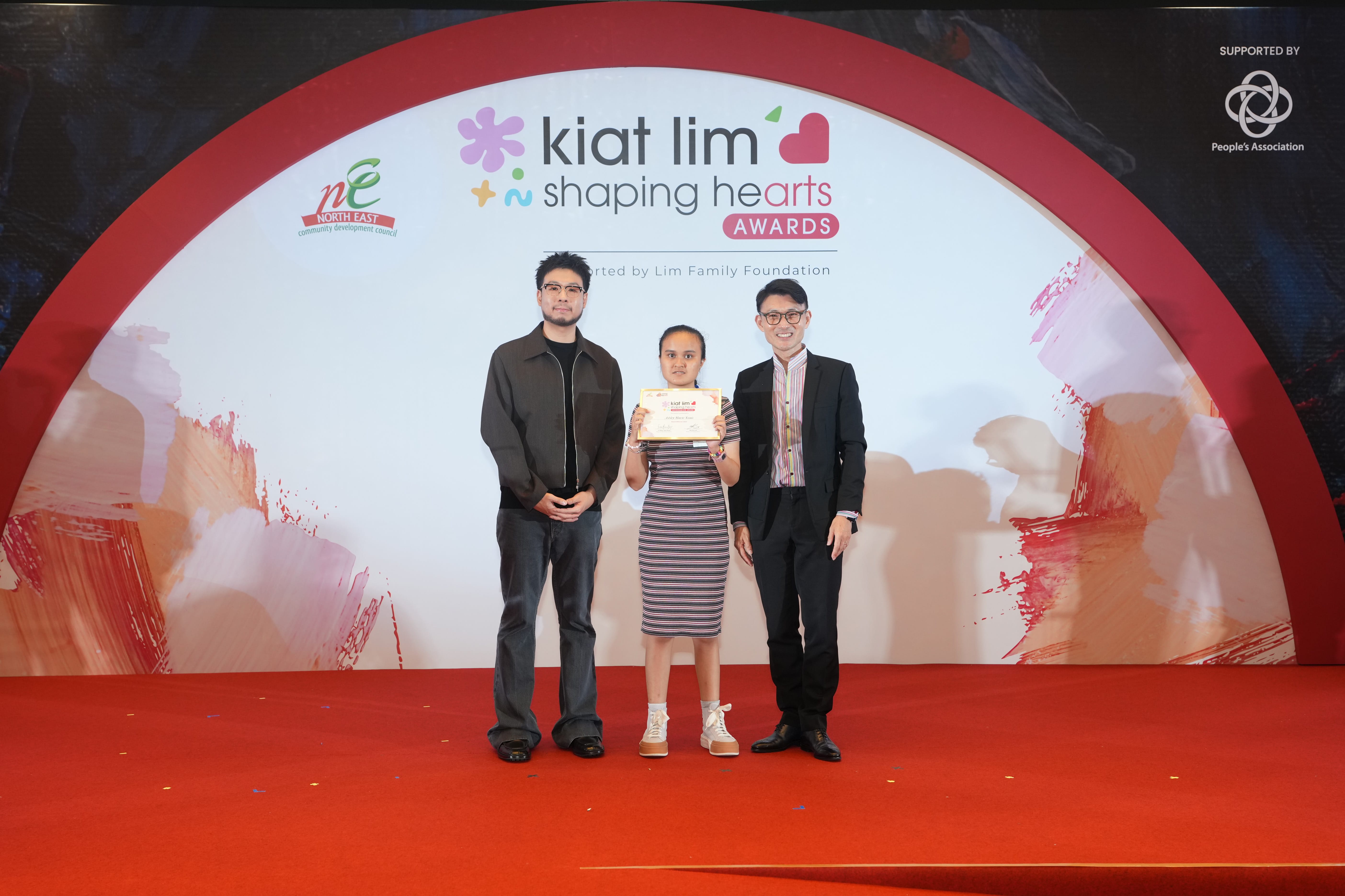 Award recipient standing on stage with the presenter, holding their certificate and posing for a group photo during the award ceremony, with a backdrop reading ‘Kiat Lim-Shaping Hearts Awards'.